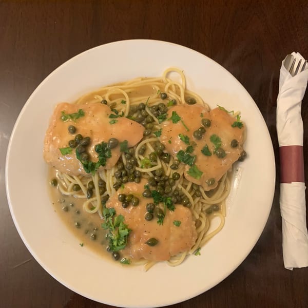 Chicken piccata with capers and parsley over spaghetti on a white plate, with a rolled napkin.