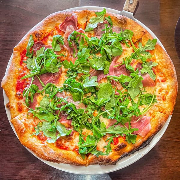 Tony's at Brentwood pizza with prosciutto, fresh arugula, and melted cheese on a white plate.